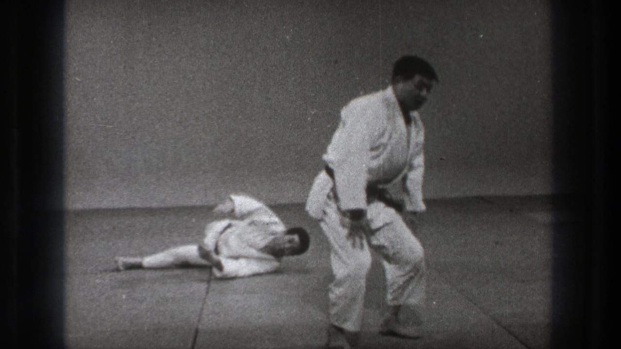 Two men in white gi on studio mats, with one standing and the other lying on the floor.