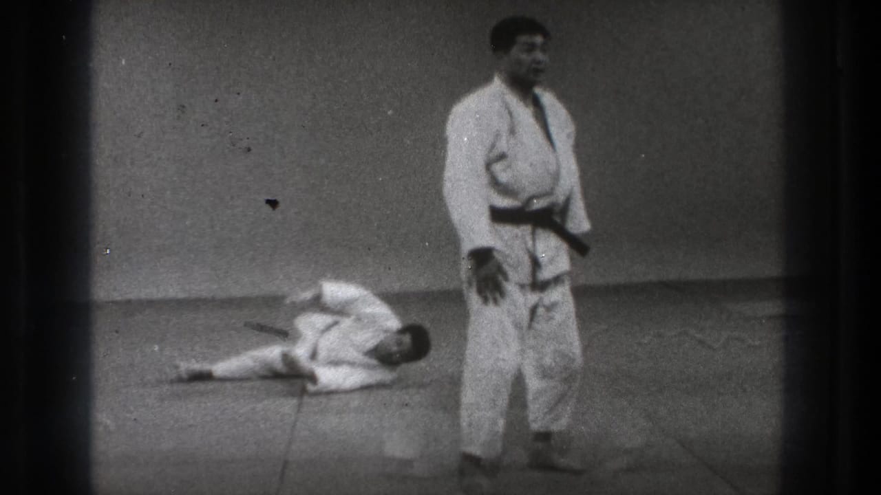 Black-and-white 8mm frame of a martial arts practitioner standing as his partner remains down on the studio mat.