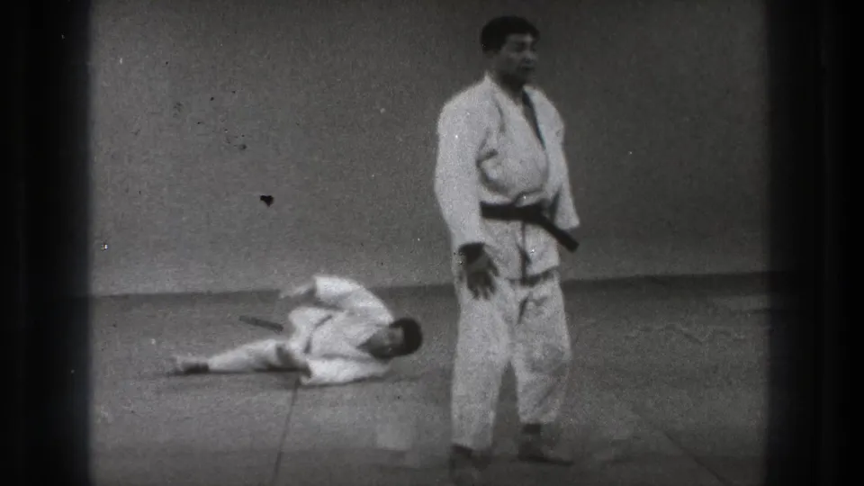 Black-and-white 8mm frame of a martial arts practitioner standing as his partner remains down on the studio mat.