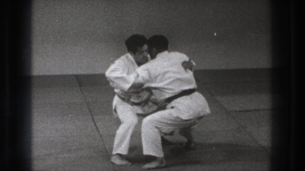 Two men in white gi crouch in close grappling stance on dojo mats.