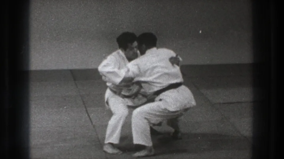 In a 1971 Tokyo dojo setting, two adult men in white gi and black belts crouch while gripping each other during practice.