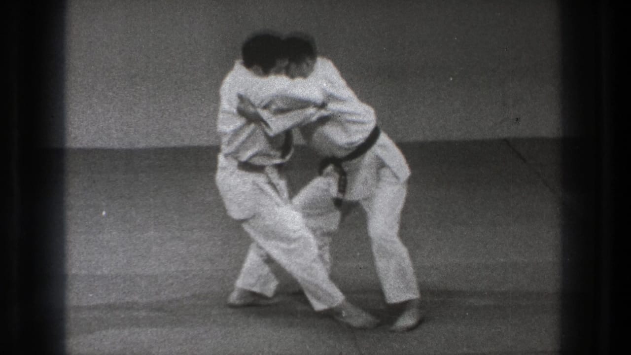 Two adult practitioners in white gi and black belts stand chest to chest in a close hold on mats in Tokyo archival footage.