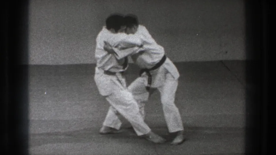 Two adult practitioners in white gi and black belts stand chest to chest in a close hold on mats in Tokyo archival footage.