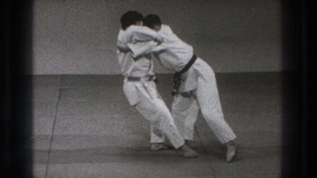 Two adult men in white gi and black belts lean into each other during a throw or balance drill on dojo mats in 1971 Tokyo footage.