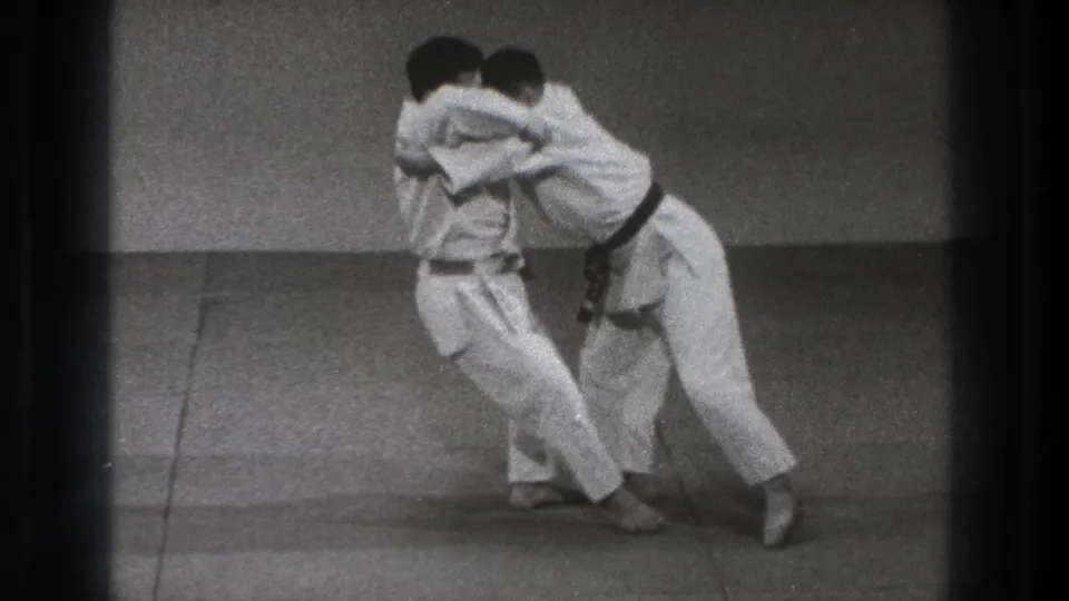 Two adult men in white gi and black belts lean into each other during a throw or balance drill on dojo mats in 1971 Tokyo footage.