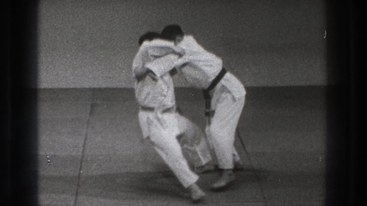 Two adult martial-arts practitioners in white gi and black belts continue a close grappling drill on mats in the Tokyo dojo footage.