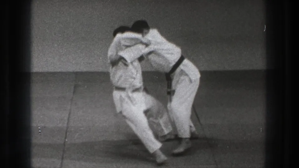 Two adult martial-arts practitioners in white gi and black belts continue a close grappling drill on mats in the Tokyo dojo footage.