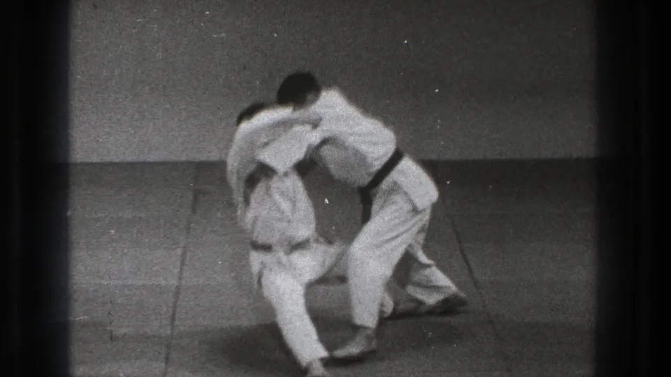 In a 1971 Tokyo martial arts setting, two men in white uniforms and dark belts grapple upright on a mat before a throw.