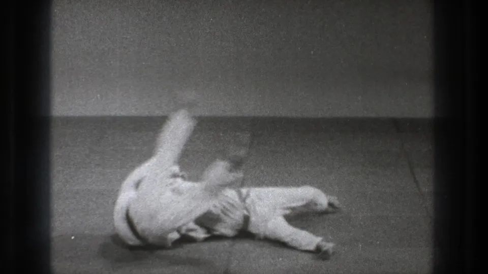 Both practitioners roll across the mat after impact, their white uniforms blurred by slow-motion movement and film grain.