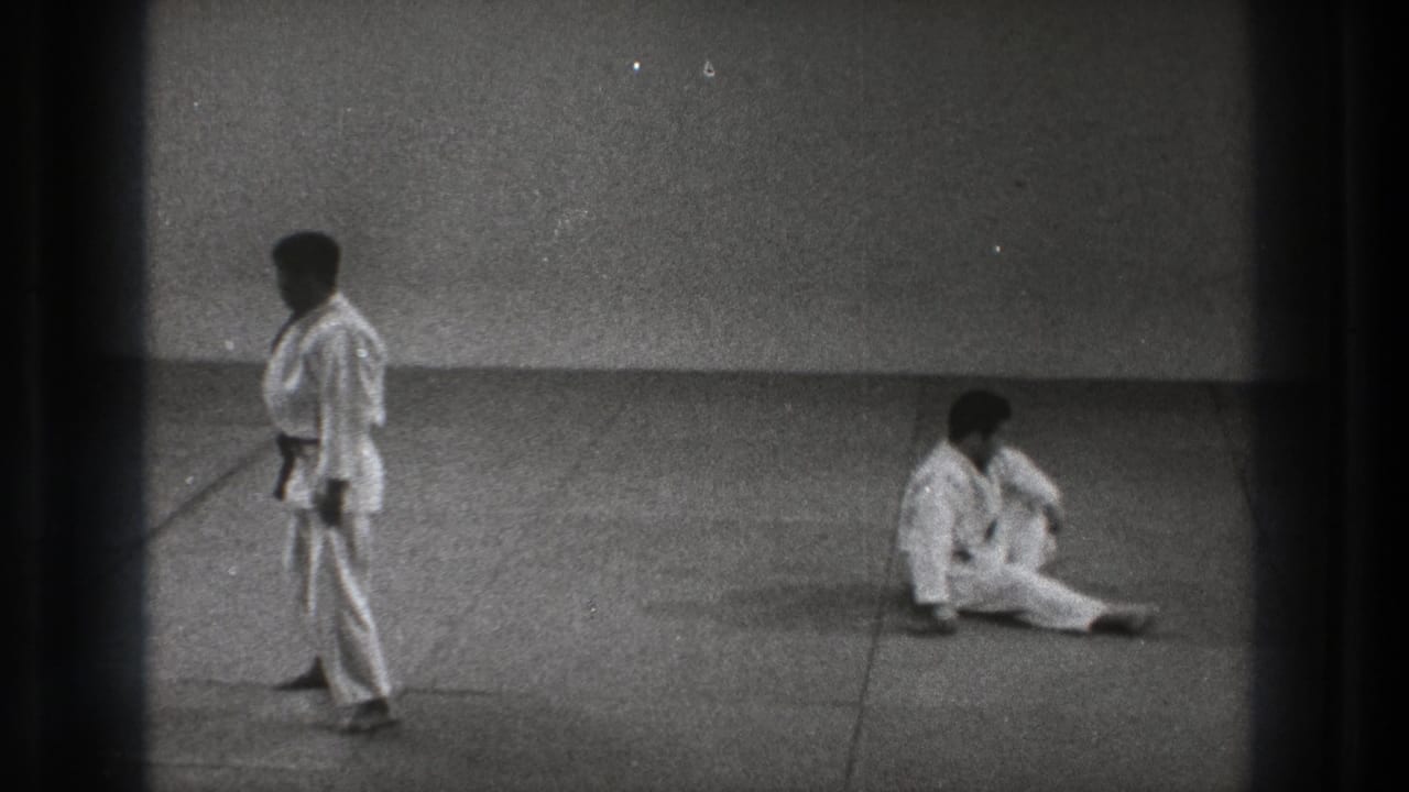In the 1971 Tokyo martial arts practice scene, one man stands in a white gi while his partner sits up on the mat after a throw.