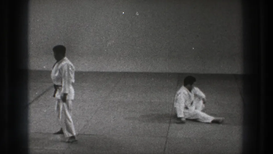 In the 1971 Tokyo martial arts practice scene, one man stands in a white gi while his partner sits up on the mat after a throw.