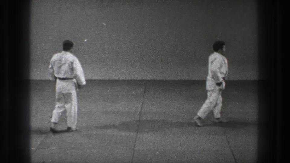 In Tokyo 1971 home-movie footage, two men in white karate uniforms stand apart on a grey mat in a plain dojo-like room.