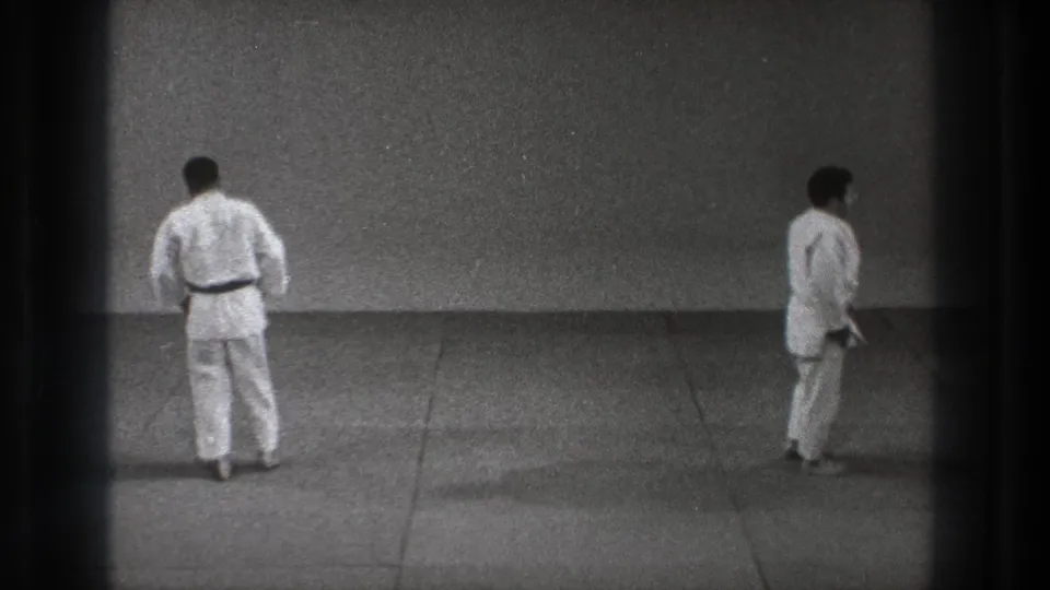 A 1971 Tokyo frame shows two men in white martial arts uniforms standing apart on a grey practice mat, framed by dark 8mm borders.
