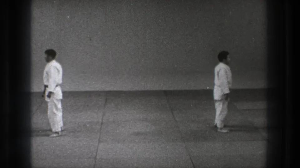 A black-and-white 8mm frame from Tokyo, 1971 shows two men in white karate uniforms standing opposite each other on a simple mat floor.