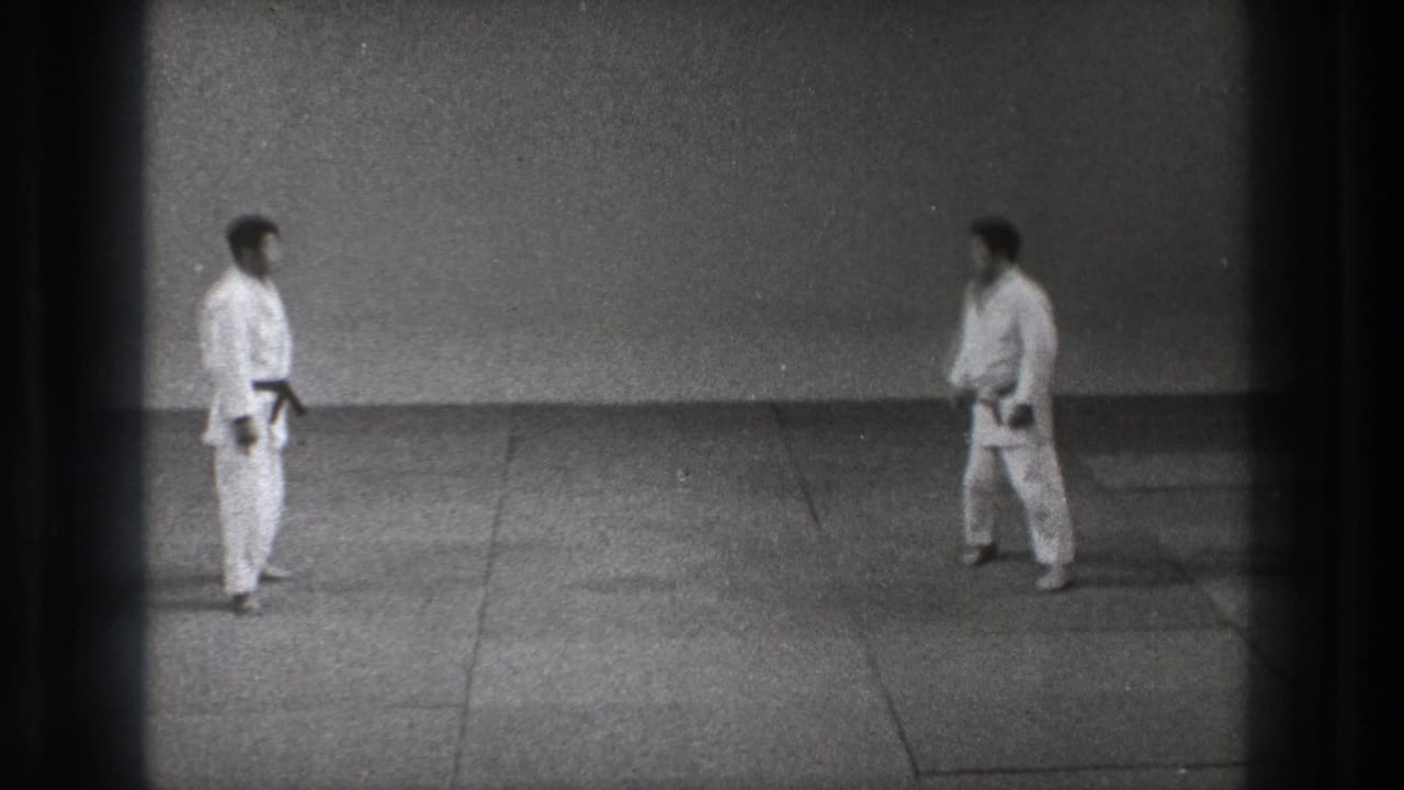 Two men in white gi and dark belts face one another in a sparse Tokyo training space, filmed on black-and-white 8mm.