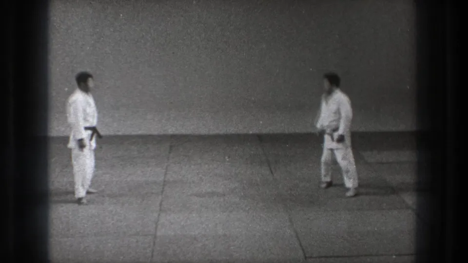 Two men in white gi and dark belts face one another in a sparse Tokyo training space, filmed on black-and-white 8mm.