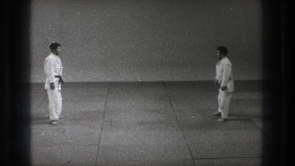 A wide 8mm frame shows two martial arts practitioners in gi maintaining distance on the mat in Tokyo, 1971.