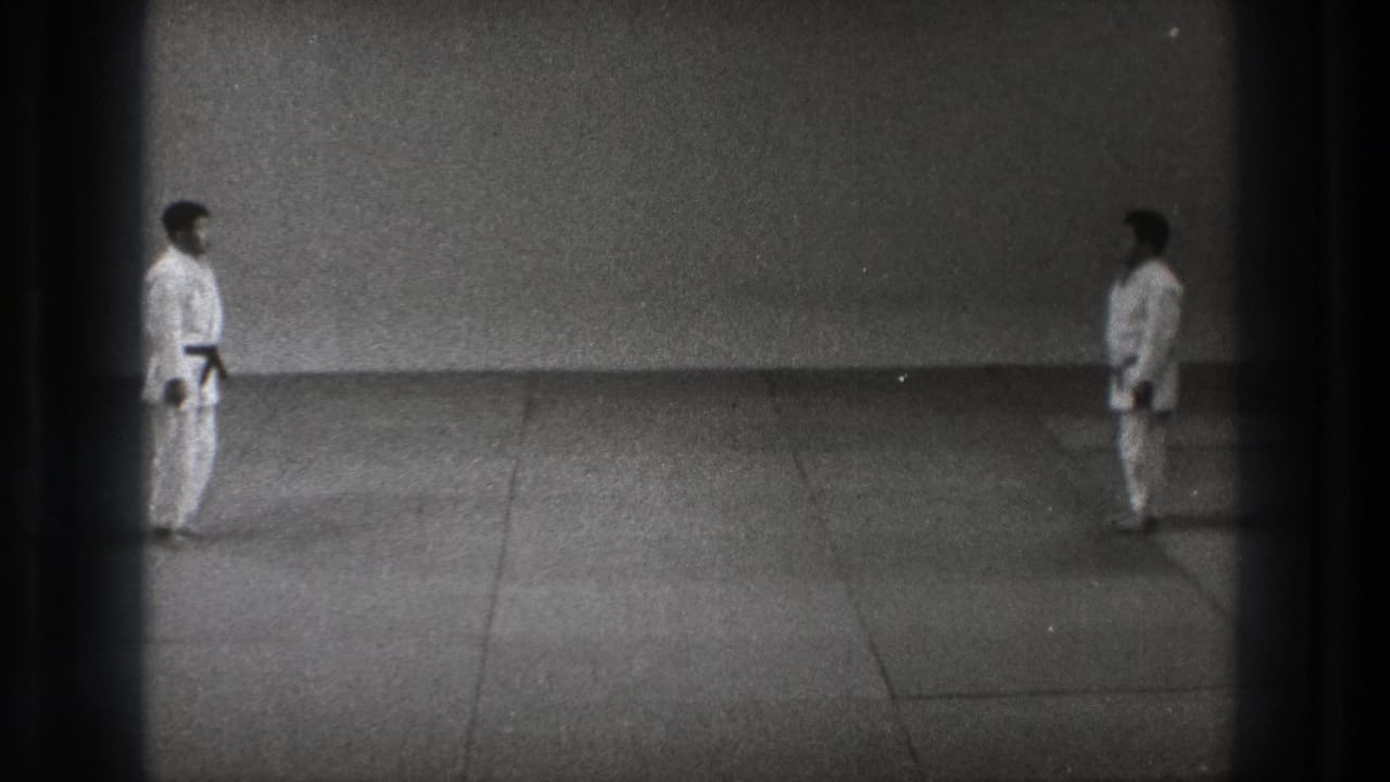 Two men in white gi remain at opposite sides of a simple training mat in black-and-white 1971 Tokyo home-movie footage.