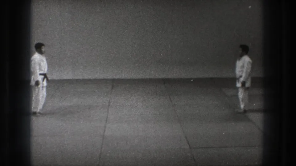 Two men in white gi remain at opposite sides of a simple training mat in black-and-white 1971 Tokyo home-movie footage.