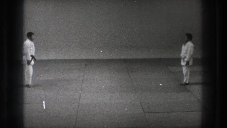 In 1971 Tokyo footage, two karate practitioners in white uniforms stand at opposite ends of a sparse mat area.