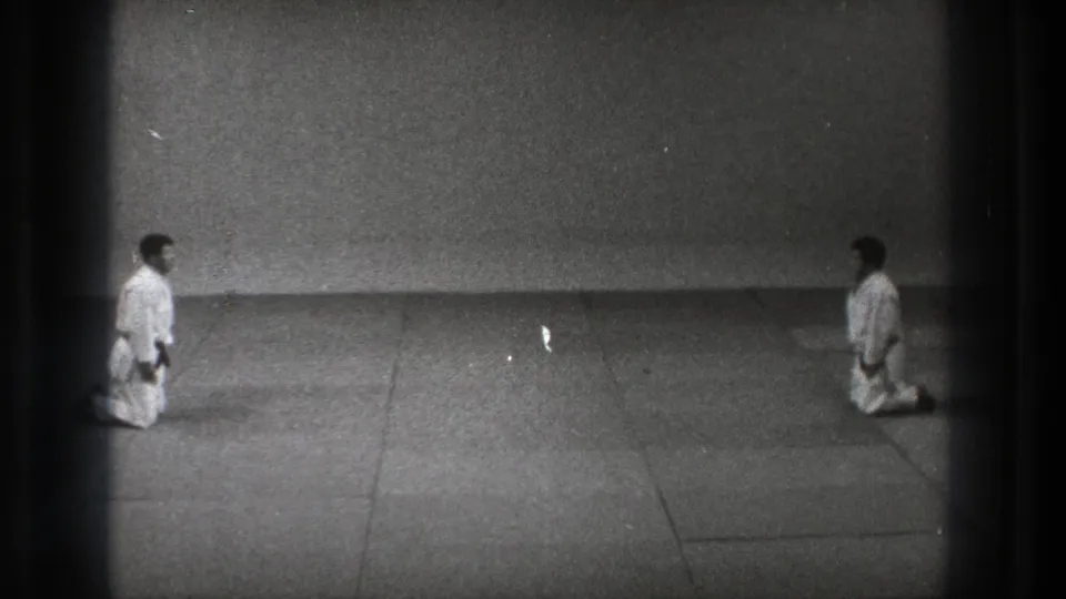The 8mm frame shows two martial artists kneeling in white uniforms, facing one another across a plain mat in Tokyo.