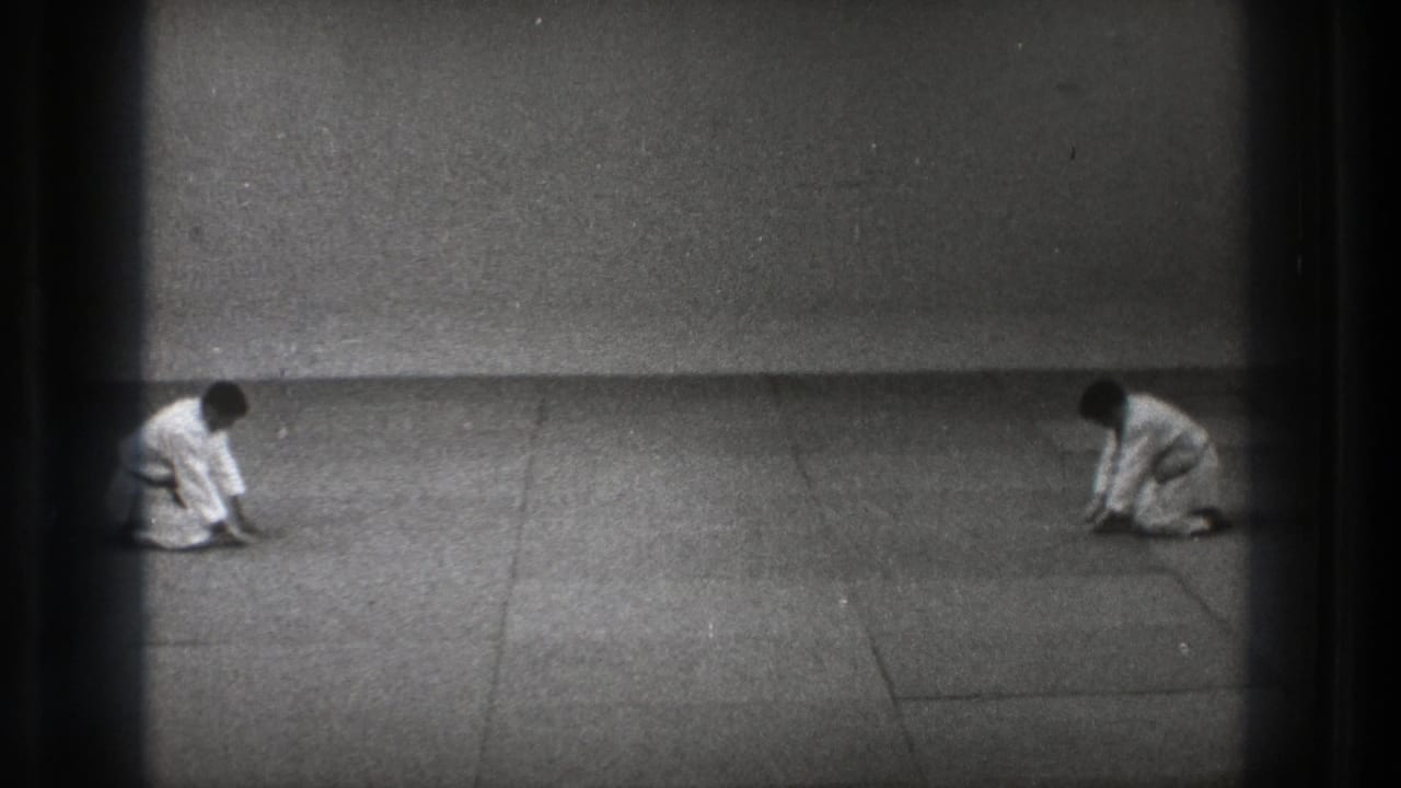 Black-and-white 8mm frame from Tokyo showing two uniformed practitioners kneeling low on a dojo mat after a bow.