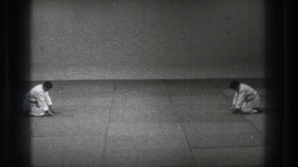 Black-and-white 8mm frame from Tokyo showing two uniformed practitioners kneeling low on a dojo mat after a bow.