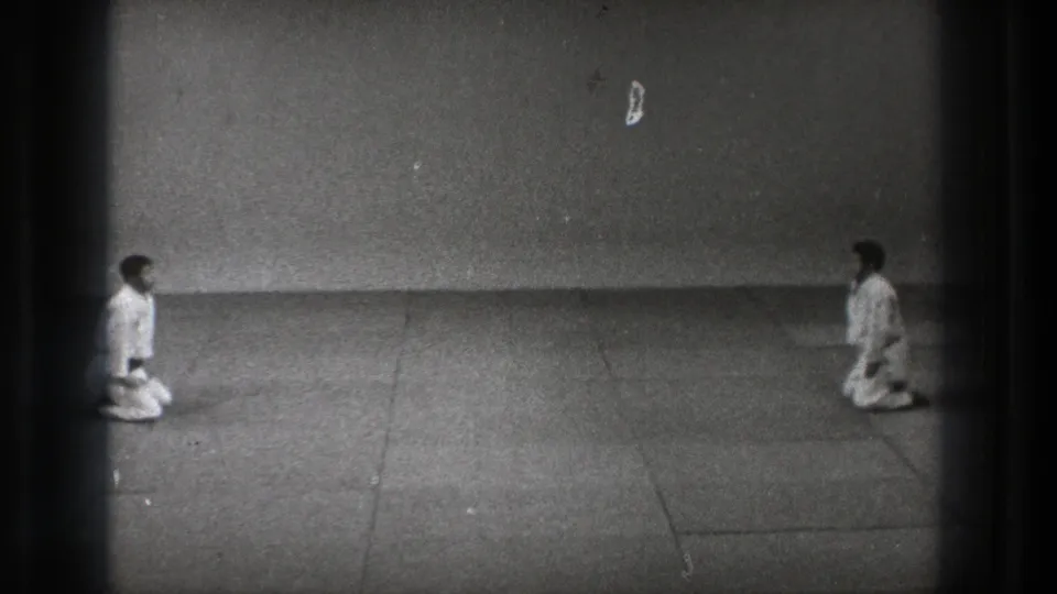 In 1971 Tokyo home-movie footage, two karate practitioners kneel at a distance on a plain mat before a bowing ritual.