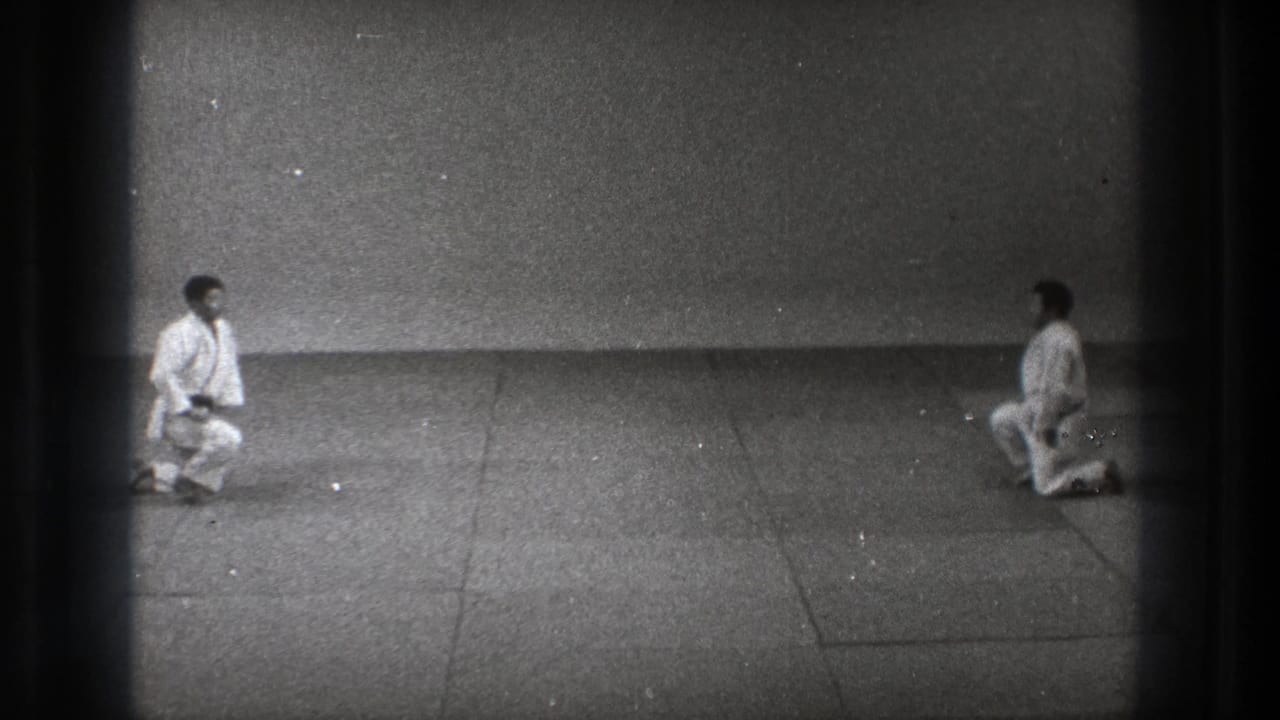 Two men in karate uniforms face each other on a mat in Tokyo, with one shifting upward from a kneeling position.