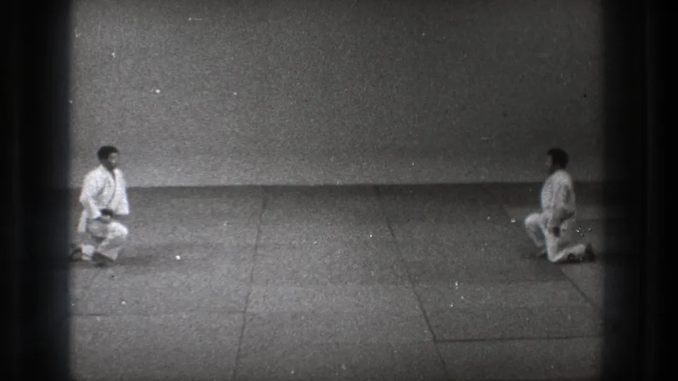 Two men in karate uniforms face each other on a mat in Tokyo, with one shifting upward from a kneeling position.