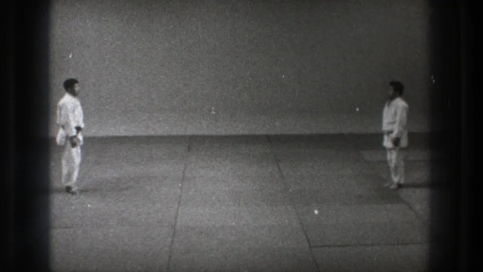 The practitioners stand apart in white gi and dark belts, facing each other in the 1971 Tokyo karate sequence.