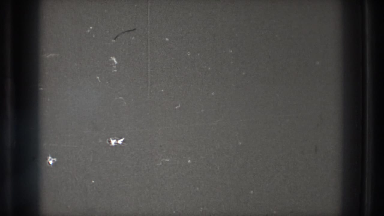 Close view of gray 8mm film material with a dark vertical edge, small dust marks, scratches, and uneven exposure.