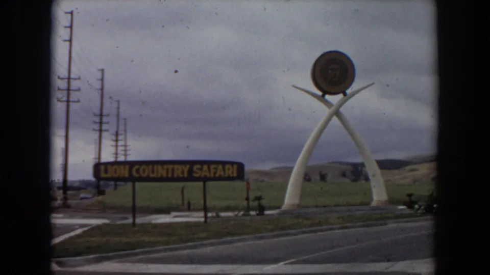 Late clip frame of the 1974 Irvine Lion Country Safari entrance, with the sign, arched sculpture, circular emblem, road foreground, and cloudy hills.