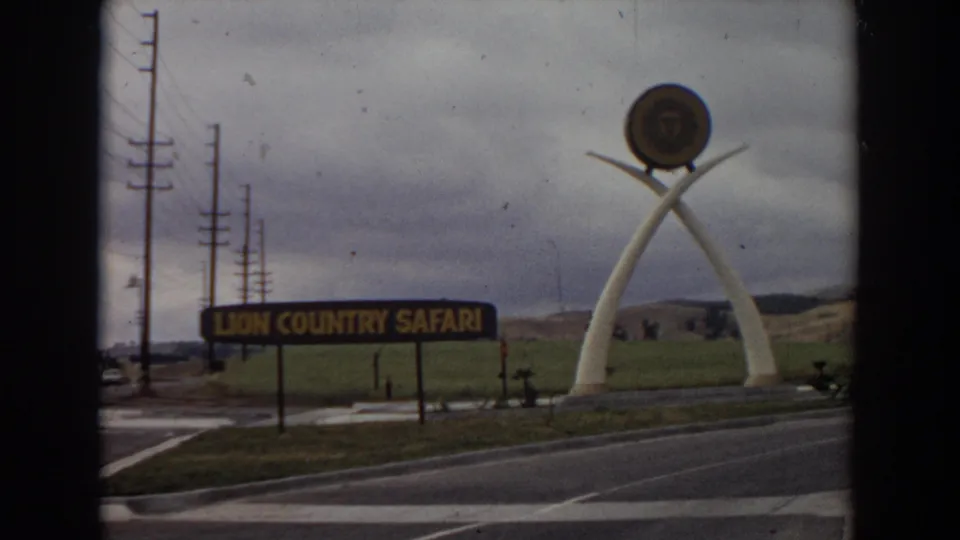 1974 Irvine, California frame showing the Lion Country Safari entrance sign, landmark white arches, circular lion emblem, road markings, utility poles, and cloudy sky.