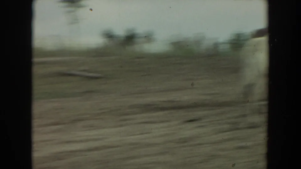 Motion-blurred 1974 Irvine 8mm view of dirt ground, low vegetation, and distant trees during a quick pan across the enclosure.