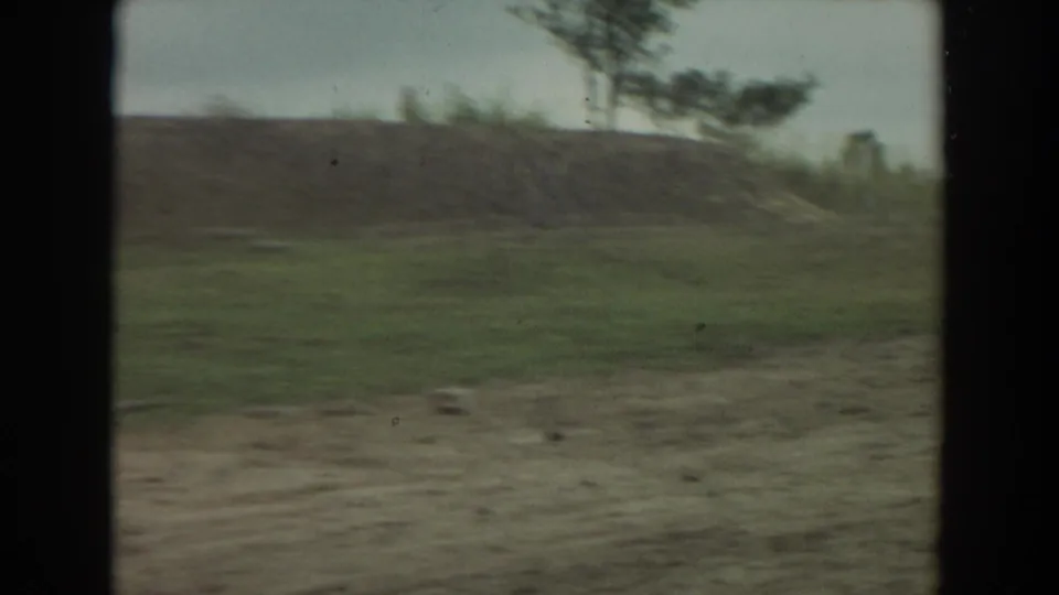 1974 Irvine 8mm frame of a grassy and dirt enclosure area with sparse trees and soft motion blur from handheld filming.
