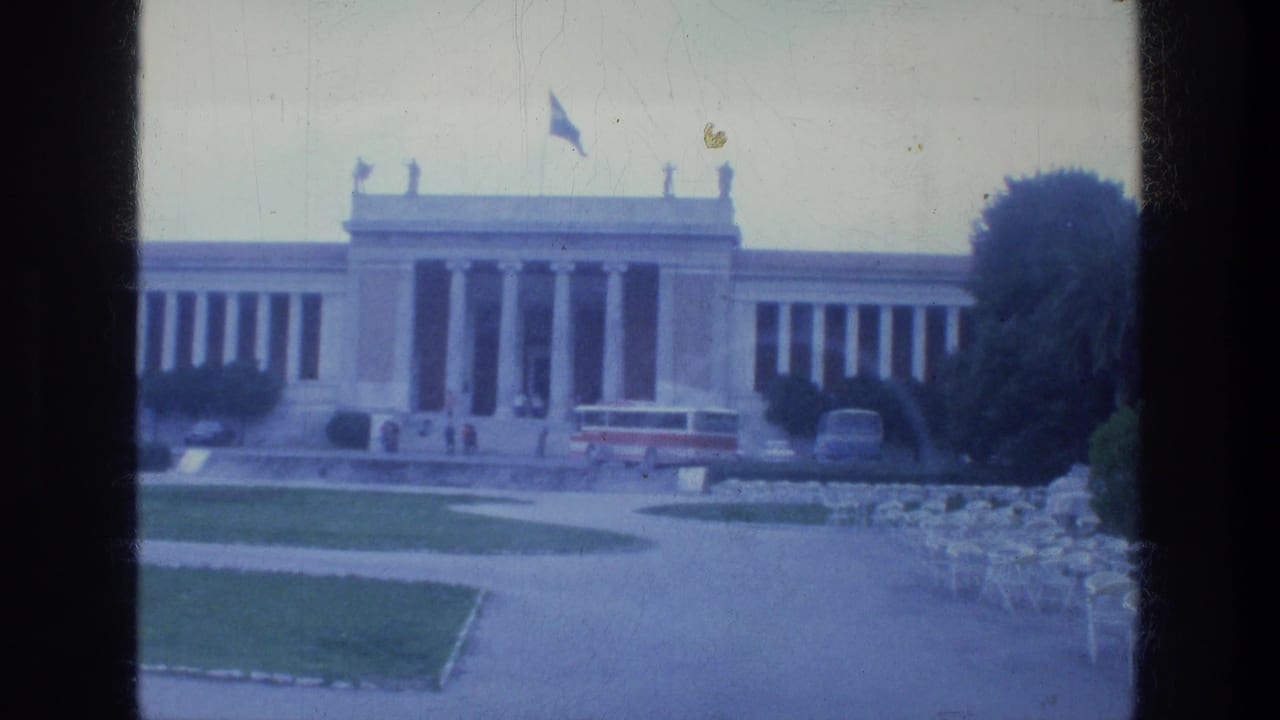 Neoclassical building with columns, Greek flag, bus, lawn, and rows of chairs in front.