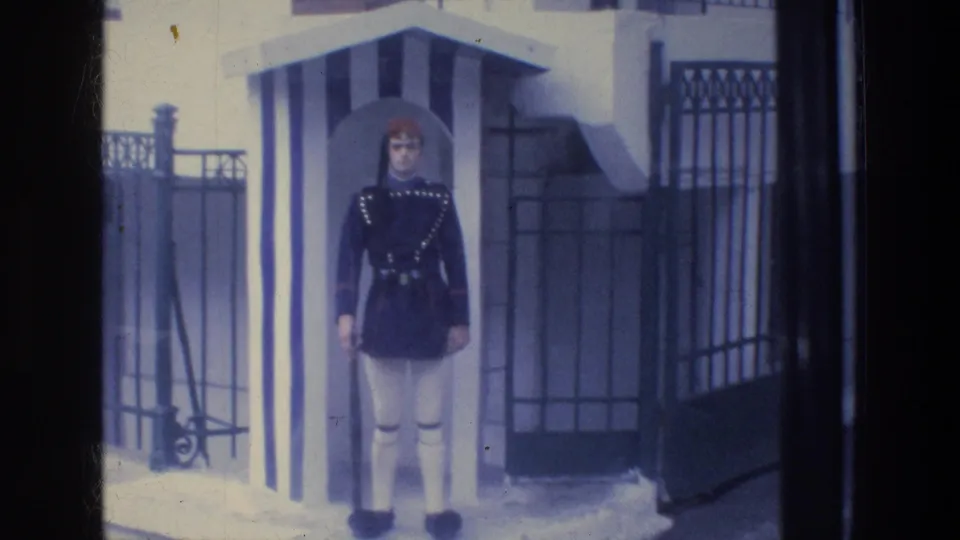 In Athens, Greece, a uniformed ceremonial guard stands at attention near a striped sentry box and iron gate in 1976 8mm footage.