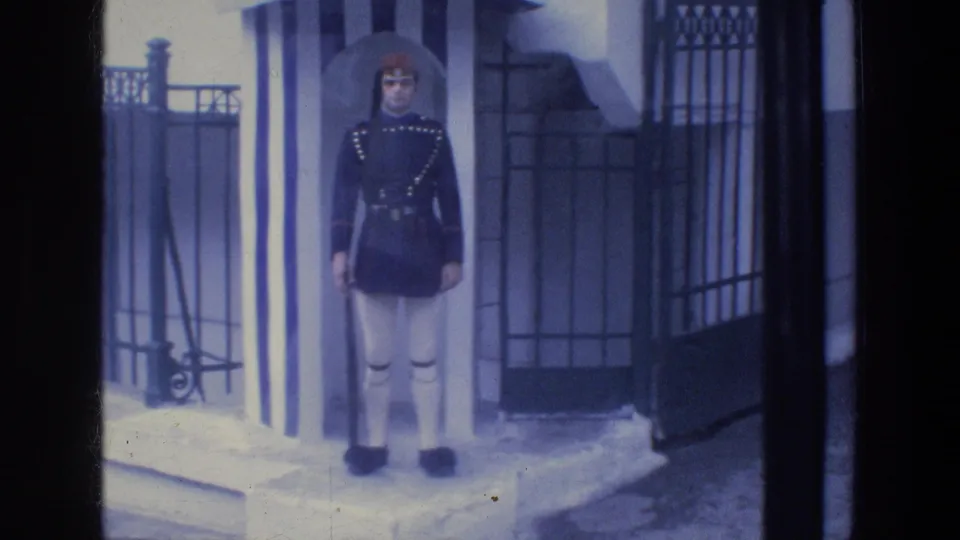 The 1976 Athens home-movie frame holds on a uniformed guard at attention before a striped sentry box and iron fence.
