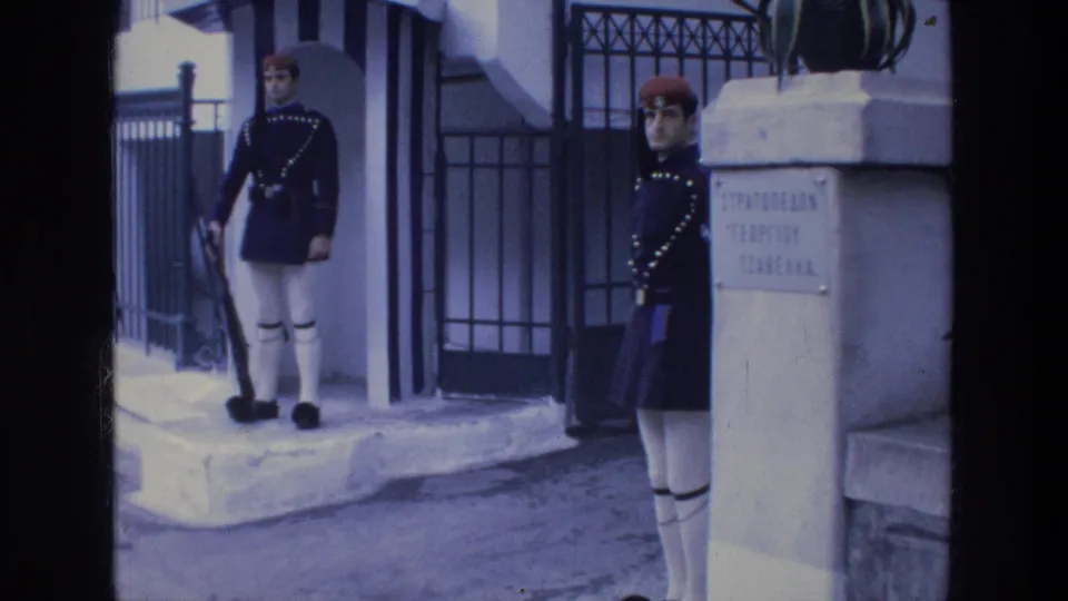 Two ceremonial guards are visible near an Athens entrance, one by the sentry box and one beside a stone pillar with Greek text.