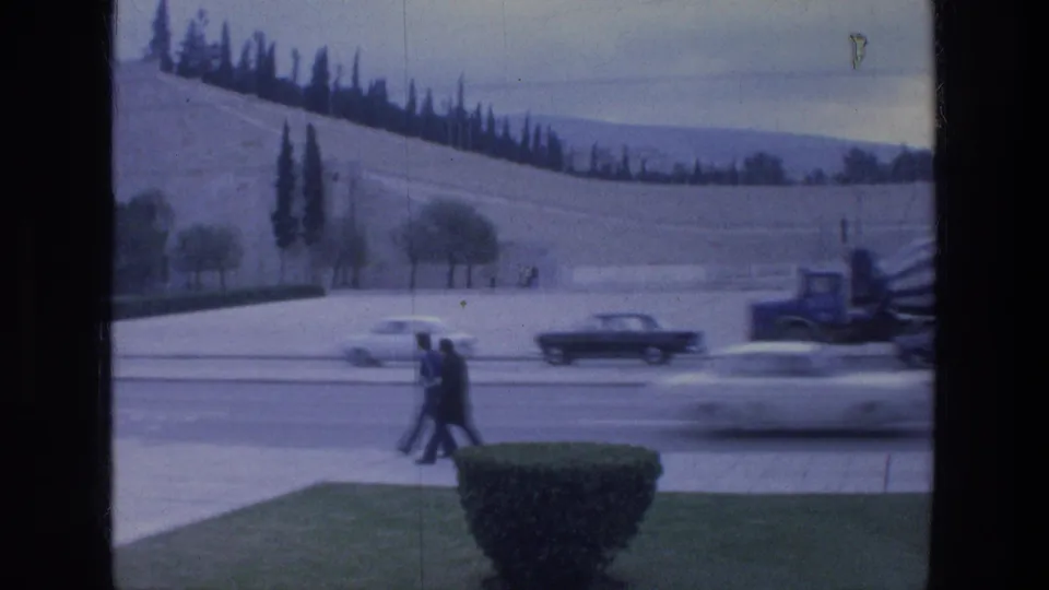 Athens 1976 street view through a window frame, with cars moving past a hillside and cypress-like trees.