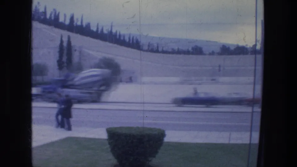 Athens 1976 home-movie view of passing cars on a busy road, with pedestrians and a hillside beyond.