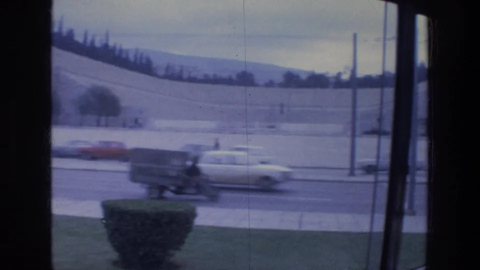 Athens 1976 traffic frame with a cement mixer truck, cars, a roadside shrub, and a distant hillside.