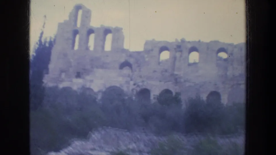 Soft 8mm view of Athens stone ruins with multiple arched openings, low greenery, and pale overcast sky.