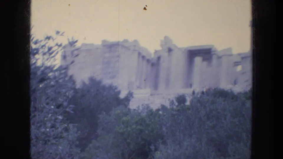 1976 8mm view of Athens ruins with stone columns and walls rising above dark trees beneath a pale sky.