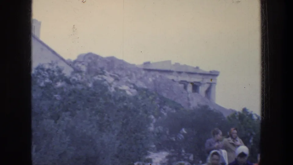 1976 Athens home-movie frame showing the Parthenon above a rocky slope with tourists near the Acropolis foreground.