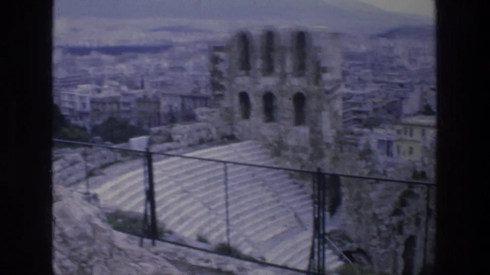 1976 Athens frame showing tan stone ruins behind chain-link fencing, with city buildings and hills beyond.