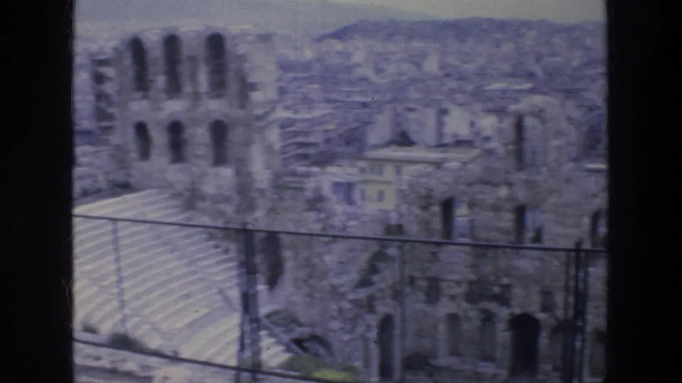 Handheld 1976 Athens view of stone ruins and chain-link fencing, with a soft city panorama in the background.