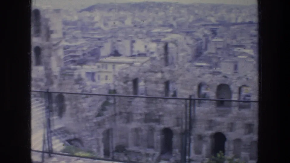 1976 Athens travel footage frame showing weathered stone arches and dense city buildings beyond, viewed with soft 8mm focus.