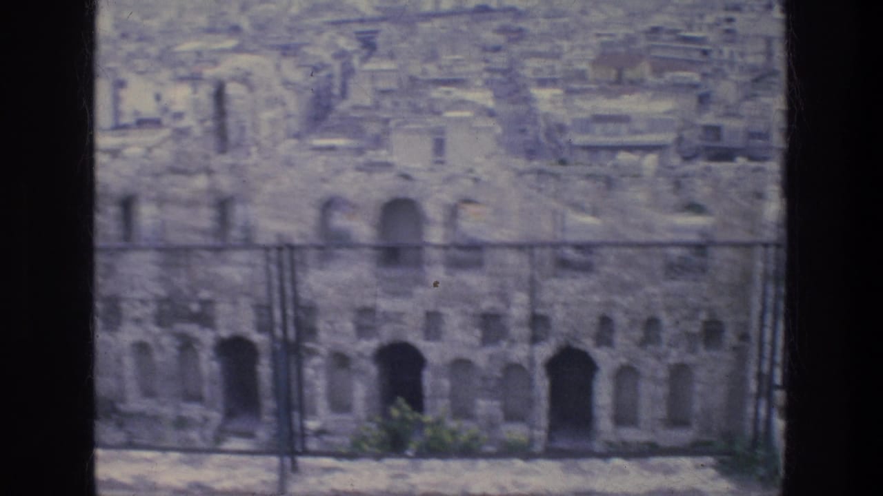 Athens ruins with arched openings and a metal barrier in front, with modern buildings filling the background.